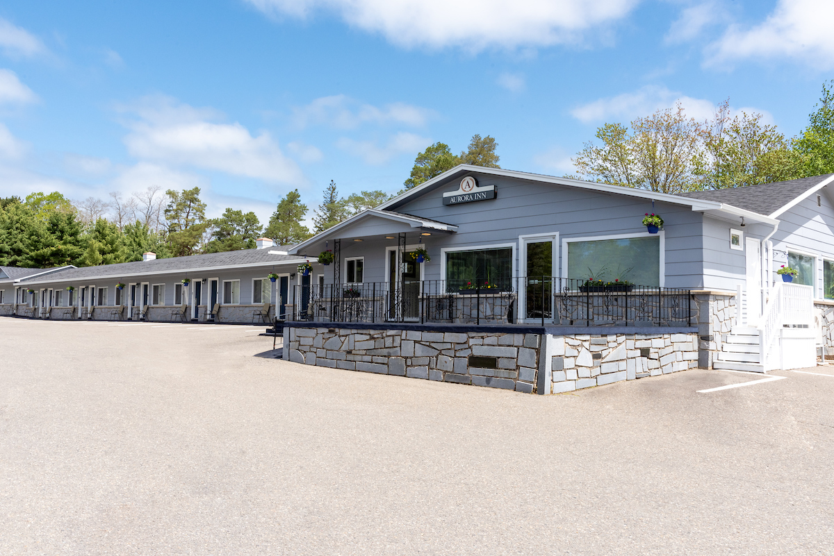 Front view of Aurora Inn motel in the Annapolis Valley showing the main entrance and guest rooms with stone accents and hanging flower baskets under a clear blue sky.